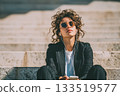 A young woman in a modern business suit sits on the steps of a building with headphones in her ears and enjoys music 133519577