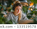 A young woman wearing headphones sits at a laptop in a modern office with lots of green plants 133519578