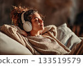 Young woman with dark curly hair wearing big white headphones listening to music with her eyes closed while sitting on a comfortable sofa at home 133519579