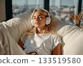 An attractive blonde with her eyes closed and wearing large white headphones listens to music while lying on a sofa in a cozy living room 133519580