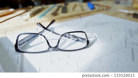 Resting black rectangular eyeglasses open on white paper at workspace, with blurred drawing tools 133520015