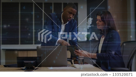 Leaning suited businessman pointing at tablet held by colleague in conference room, with laptop 133520071