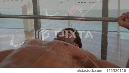 Lifting shirtless mid adult male bench pressing in gym, with barbell and floor-to-ceiling windows 133520116
