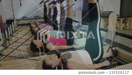 Lifting legs by six women on Pilates reformers in fitness studio, showing foot bars and springs 133520117