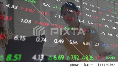 Coworkers reviewing financial data overlay on office desk with laptop, one wearing mustard dress 133520162