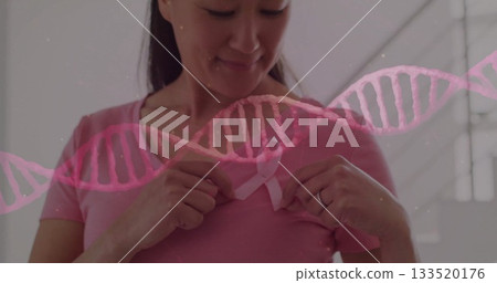 Korean woman fastening awareness ribbon over pink t-shirt in softly lit room, with DNA overlay Korean woman fastening awareness ribbon over pink t-shirt in softly lit room, with DNA overlay 133520176
