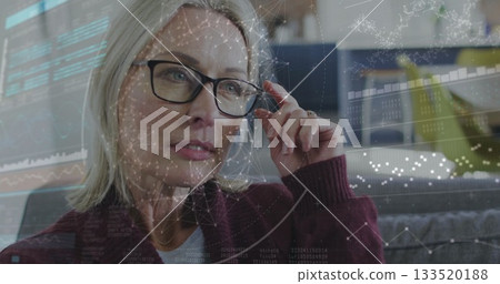 Woman adjusting black glasses while analyzing digital overlays on sofa wearing burgundy cardigan 133520188
