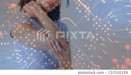 Standing woman wearing blue shirt holding right elbow inside room, with digital circuit overlay 133520189