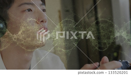 Sitting Korean teen wearing over-ear headphones and holding gamepad in room, with network overlay 133520201