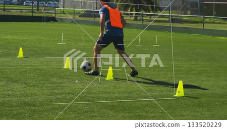 Soccer player wearing orange vest dribbling ball through yellow cones on soccer pitch, with poles 133520229