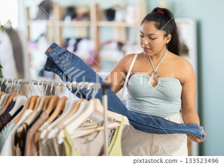 Positive young woman chooses fashionable jeans in clothing store 133523496
