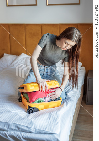 Young woman struggling with overstuffed suitcase on bed Young woman struggling with overstuffed suitcase on bed 133527982