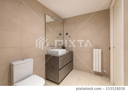 Modern bathroom with beige tiles, a white toilet, a sleek sink, and a large mirror. Minimalist design with a radiator and natural light. 133528038