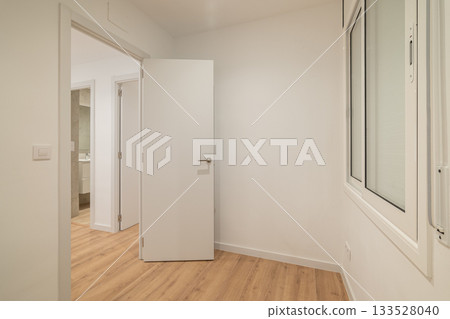 A modern, empty room with white walls and wooden flooring. A closed white door is visible, leading to another space. Natural light enters through a window. 133528040