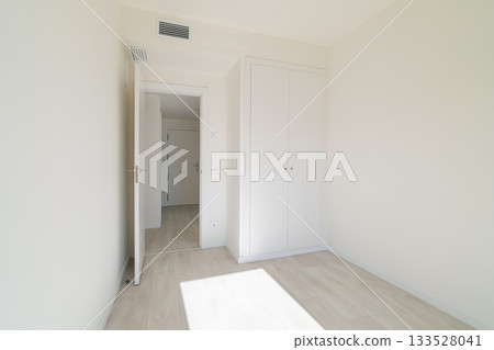 A bright, empty room with white walls and light wooden flooring. A closed door leads to another space, and a wardrobe is visible against the wall. 133528041