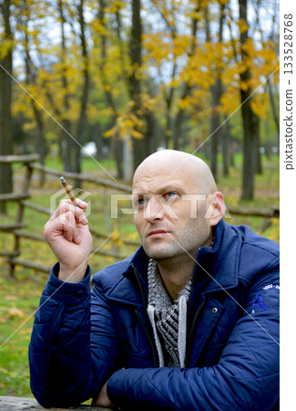 bald young man in a jacket smokes a cigarette on a bench in the autumn park 133528768