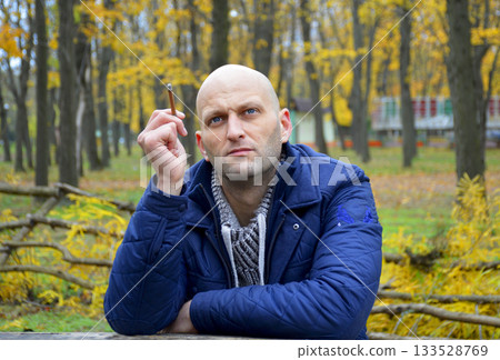 bald young man in a jacket smokes a cigarette on a bench in the autumn park bald young man in a jacket smokes a cigarette on a bench in the autumn park 133528769