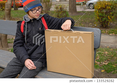 boy sits on a bench holding a box by the handle looks down 133529282