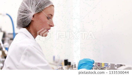 Operating technician wearing lab coat, hairnet, adjusting panel labels in lab, with vials, tubing 133530044