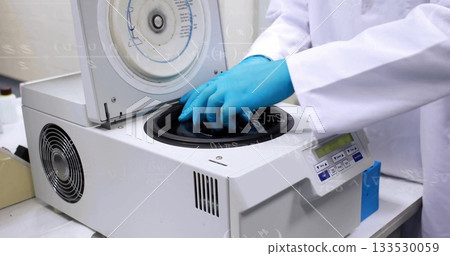 Technician wearing white lab coat loading sample tubes into benchtop centrifuge at lab bench Technician wearing white lab coat loading sample tubes into benchtop centrifuge at lab bench 133530059