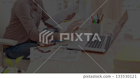Woman wearing button-down shirt holding documents while using laptop at office desk, with pencils 133530121