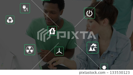 Collaborating adults pointing stylus at laptop screen in office showing floating green energy icons 133530123