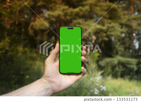 Hand holding mobile phone with empty white screen in front of blurred green forest background. for 133531173