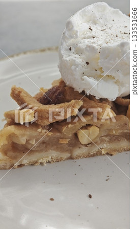 Slice of apple pie with nuts topped with a scoop of vanilla ice cream on a white plate. this image conveys sweetness, comfort and cozy dessert enjoyment 133531666
