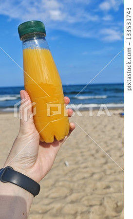 Hand holding a bottle of orange juice on a sunny beach with blue sea in the background. this image conveys refreshment and summer relaxation Hand holding a bottle of orange juice on a sunny beach with blue sea in the background. this image conveys refreshment and summer relaxation 133531743