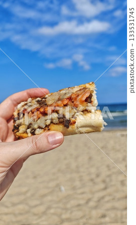 Hand holding a slice of baked bread with melted toppings against a sunny beach and blue sky. this image conveys relaxation, outdoor snack and summer leisure Hand holding a slice of baked bread with melted toppings against a sunny beach and blue sky. this image conveys relaxation, outdoor snack and summer leisure 133531745