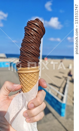 Hand holding a tall swirl of chocolate ice cream in a waffle cone against a sunny beach background. this image conveys enjoyment, summer treat and carefree leisure Hand holding a tall swirl of chocolate ice cream in a waffle cone against a sunny beach background. this image conveys enjoyment, summer treat and carefree leisure 133531747