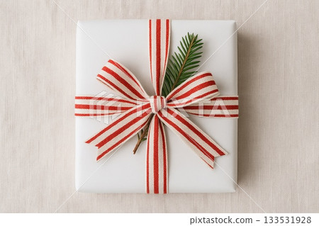 White gift box wrapped with a red striped ribbon and a small evergreen sprig on a neutral background. this image conveys holiday giving, festive simplicity and cozy seasonal charm 133531928