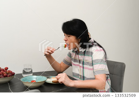 Middle aged Russian woman eating fresh vegetables at home table with simple meal, grapes and water, everyday dining moment. 133532509