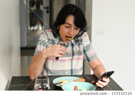 Mature 50 year old Russian woman eats breakfast while staring at her phone, showing a strong phone addiction and an unhealthy distracted habit. Mature 50 year old Russian woman eats breakfast while staring at her phone, showing a strong phone addiction and an unhealthy distracted habit. 133532510