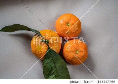 Close-up of three fresh tangerines with a green leaf on a beige background. Minimal and natural composition suitable for holiday, Christmas, New Year, or fresh fruit concepts. Close-up of three fresh tangerines with a green leaf on a beige background. Minimal and natural composition suitable for holiday, Christmas, New Year, or fresh fruit concepts. 133532866