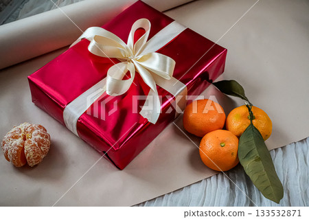 Side view of a red gift box with a ribbon placed on a beige background. Three tangerines lie beside the box, including one peeled tangerine with a small green twig. Perfect for Christmas, New Year Side view of a red gift box with a ribbon placed on a beige background. Three tangerines lie beside the box, including one peeled tangerine with a small green twig. Perfect for Christmas, New Year 133532871