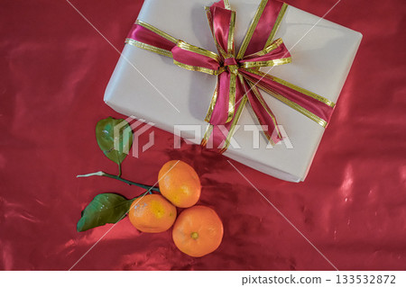 Medium shot of a beige gift box with a red ribbon placed next to three fresh tangerines on a solid red background. Festive and minimal still life composition suitable for holiday themes. Medium shot of a beige gift box with a red ribbon placed next to three fresh tangerines on a solid red background. Festive and minimal still life composition suitable for holiday themes. 133532872