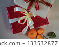 Horizontal close-up view of two stacked gift boxes, one red and one beige, decorated with ribbons. Fresh tangerines lie beside the boxes on a beige background, forming a warm and minimal holiday still 133532877