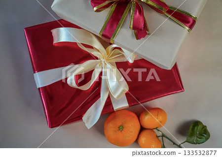 Horizontal close-up view of two stacked gift boxes, one red and one beige, decorated with ribbons. Fresh tangerines lie beside the boxes on a beige background, forming a warm and minimal holiday still Horizontal close-up view of two stacked gift boxes, one red and one beige, decorated with ribbons. Fresh tangerines lie beside the boxes on a beige background, forming a warm and minimal holiday still 133532877