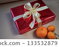 Side view of a red gift box with a ribbon bow placed on a beige background. Three fresh tangerines are arranged beside it, creating a warm and simple holiday still life. 133532879