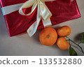 Close-up top view of a half-visible red gift box with a ribbon placed on a beige background. Three fresh tangerines with a small green twig lie beside it, creating a warm and minimal holiday still 133532880