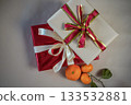 Top view of beige and red gift boxes with ribbons placed together on a neutral beige background. Three fresh tangerines with a small green twig lie nearby, creating a warm festive composition. 133532881