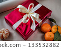 Close-up view of a red gift box with a ribbon on a beige background. Three tangerines are placed nearby, including one peeled tangerine with a small green twig. Great for holiday, Christmas, New Year 133532882