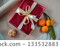 Top view of a red gift box with a ribbon placed on a beige background. Three tangerines lie beside the box, including one peeled tangerine with a small green twig, creating a festive and warm holiday 133532883