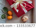 Close-up view of a half-visible red gift box with a ribbon, accompanied by fresh tangerines on a neutral background. Minimal and festive composition suitable for Christmas, New Year, holiday, or 133533677