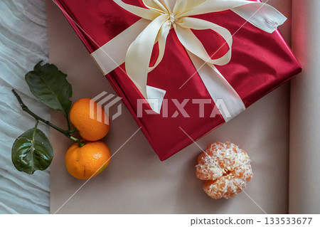 Close-up view of a half-visible red gift box with a ribbon, accompanied by fresh tangerines on a neutral background. Minimal and festive composition suitable for Christmas, New Year, holiday, or 133533677