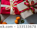 Close-up view of a red gift box with a beige gift box stacked on top, decorated with ribbons. A fresh tangerine lies on top, creating a minimal and festive composition for Christmas, New Year 133533678
