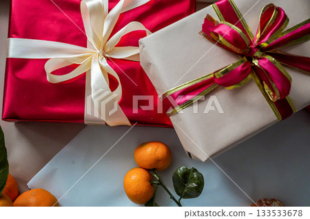 Close-up view of a red gift box with a beige gift box stacked on top, decorated with ribbons. A fresh tangerine lies on top, creating a minimal and festive composition for Christmas, New Year 133533678