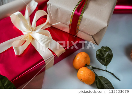 Close-up view of a red gift box with a beige gift box on top, both decorated with ribbons. A fresh tangerine lies on top, creating a warm and festive composition suitable for Christmas, New Year Close-up view of a red gift box with a beige gift box on top, both decorated with ribbons. A fresh tangerine lies on top, creating a warm and festive composition suitable for Christmas, New Year 133533679