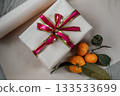 Medium view of a beige gift box decorated with a red ribbon, placed on a neutral background. Two fresh tangerines lie beside it, creating a warm and festive composition suitable for Christmas, New 133533699